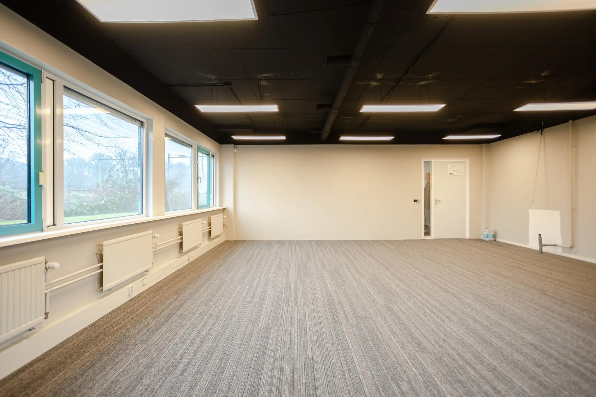 Lege kantoorruimte aan de Christiaan Krammlaan met grijs tapijt, grote ramen en zwarte systeemplafonds.